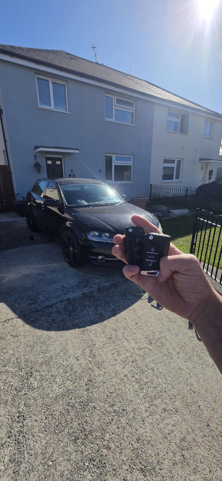 Two Audi key fobs held in front of a black Audi estate on a driveway