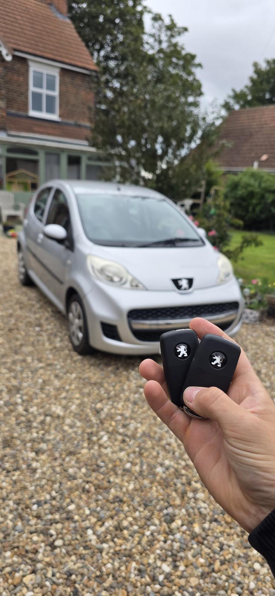 Two Peugeot key fobs held in front of a silver Peugeot on a gravel drive
